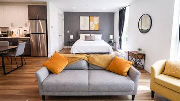 Une chambre d’hôtel moderne avec un canapé gris, un fauteuil jaune, un lit avec une literie blanche, un coin cuisine et un mur d’accent gris agrémenté d’œuvres d’art.