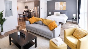 Una sala de estar moderna con un sofá gris, un sillón amarillo, una mesa de centro negra y una cama con ropa de cama blanca.