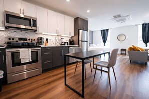 Una cucina moderna con elettrodomestici in acciaio inossidabile, una zona pranzo con tavolo e sedie, e un soggiorno con divano e una finestra.