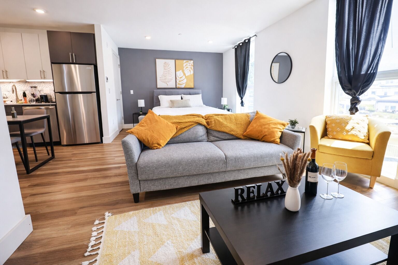 Un salon moderne avec un canapé gris, des coussins jaunes, une table basse noire sur laquelle se trouvent une bouteille et des verres, et une cuisine en arrière-plan.