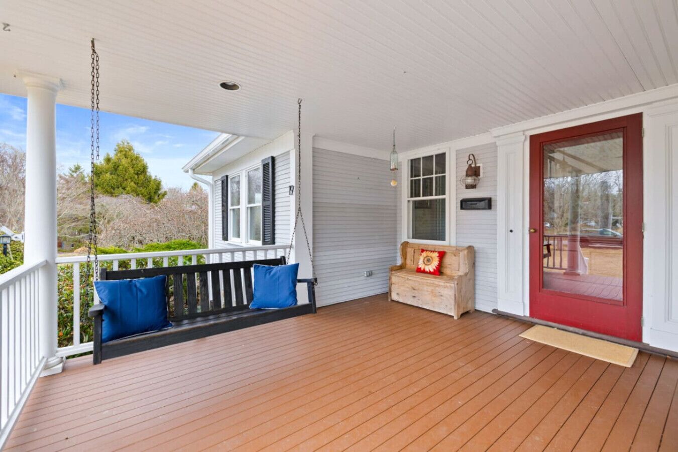 A wooden porch with a swing, a bench with cushions, a small wooden chest, and a red door.