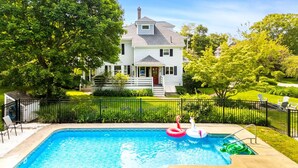 Une piscine rectangulaire entourée d’une clôture en métal noir, elle-même bordée d’une pelouse bien entretenue et d’une maison blanche dotée d’un porche.