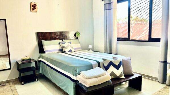 A neatly made bed with striped bedding and multiple pillows, a bedside table with a lamp, and a window with curtains.