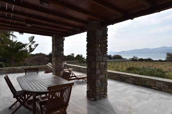 An outdoor patio with a stone column structure, a wooden table with chairs, and a view of a body of water and mountains in the distance. - Cycladic home in Kampos by GHH (Paros)