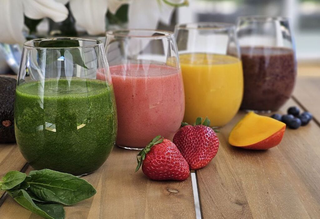 A wooden table with four glasses of smoothies and fresh fruits like strawberries and mango.