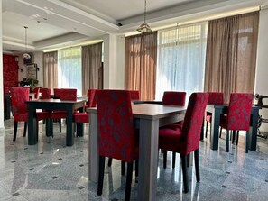 A dining area with red upholstered chairs and tables, large windows with curtains, and a polished tile floor. - West Gate (Sutomore)