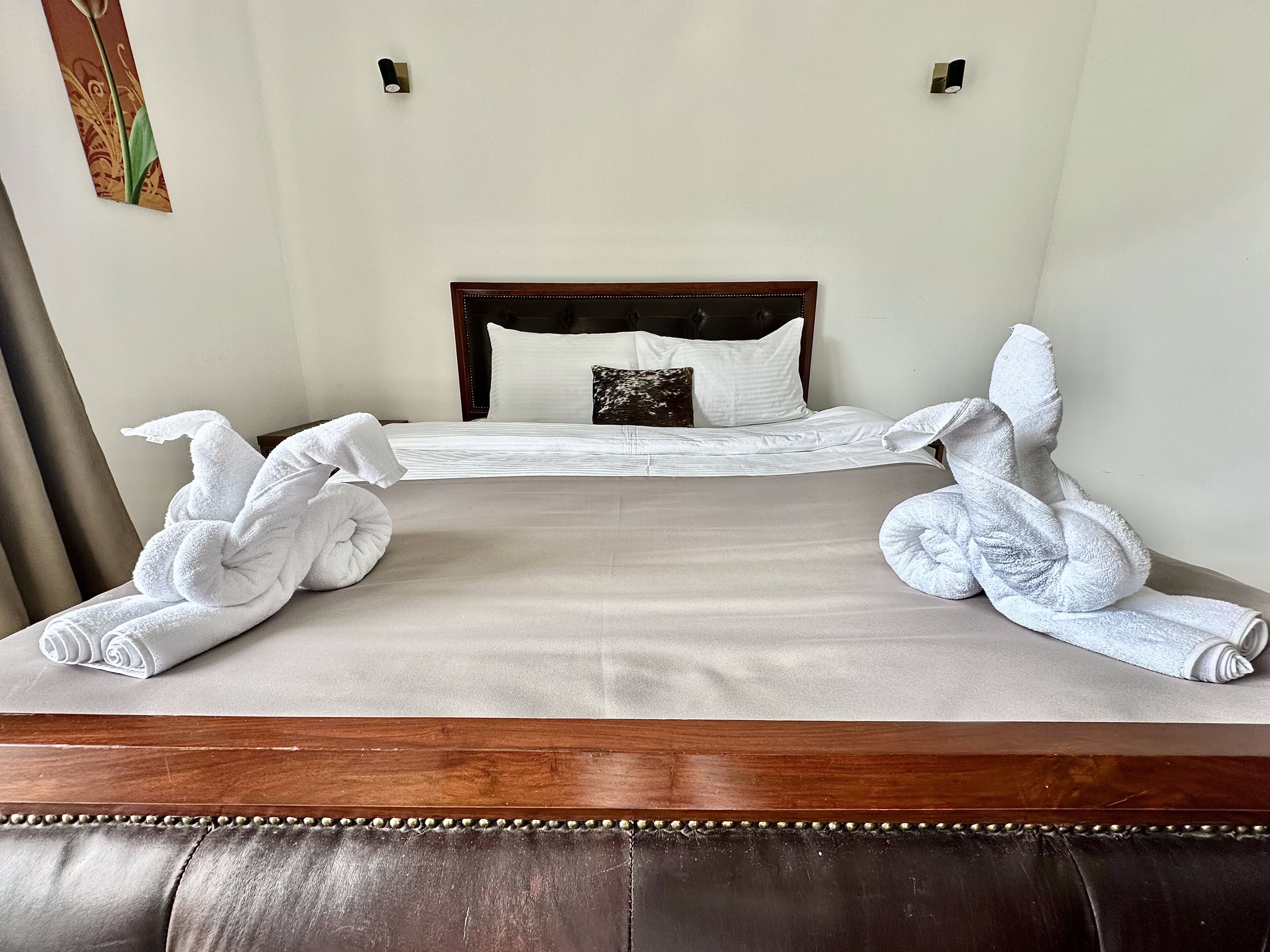 A neatly made bed with white towels arranged in a decorative pattern, a wooden headboard, and a dark leather ottoman.