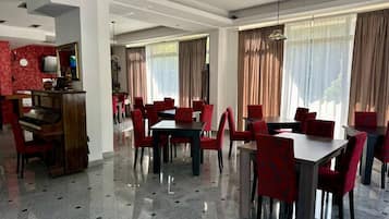 Un comedor de hotel con varias mesas y sillas, un piano y ventanas grandes con cortinas.