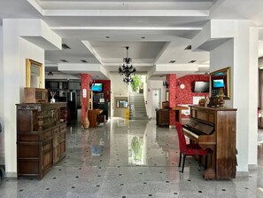 A hotel lobby with a grand piano, a reception desk, and a staircase leading to upper floors. - West Gate (Sutomore)