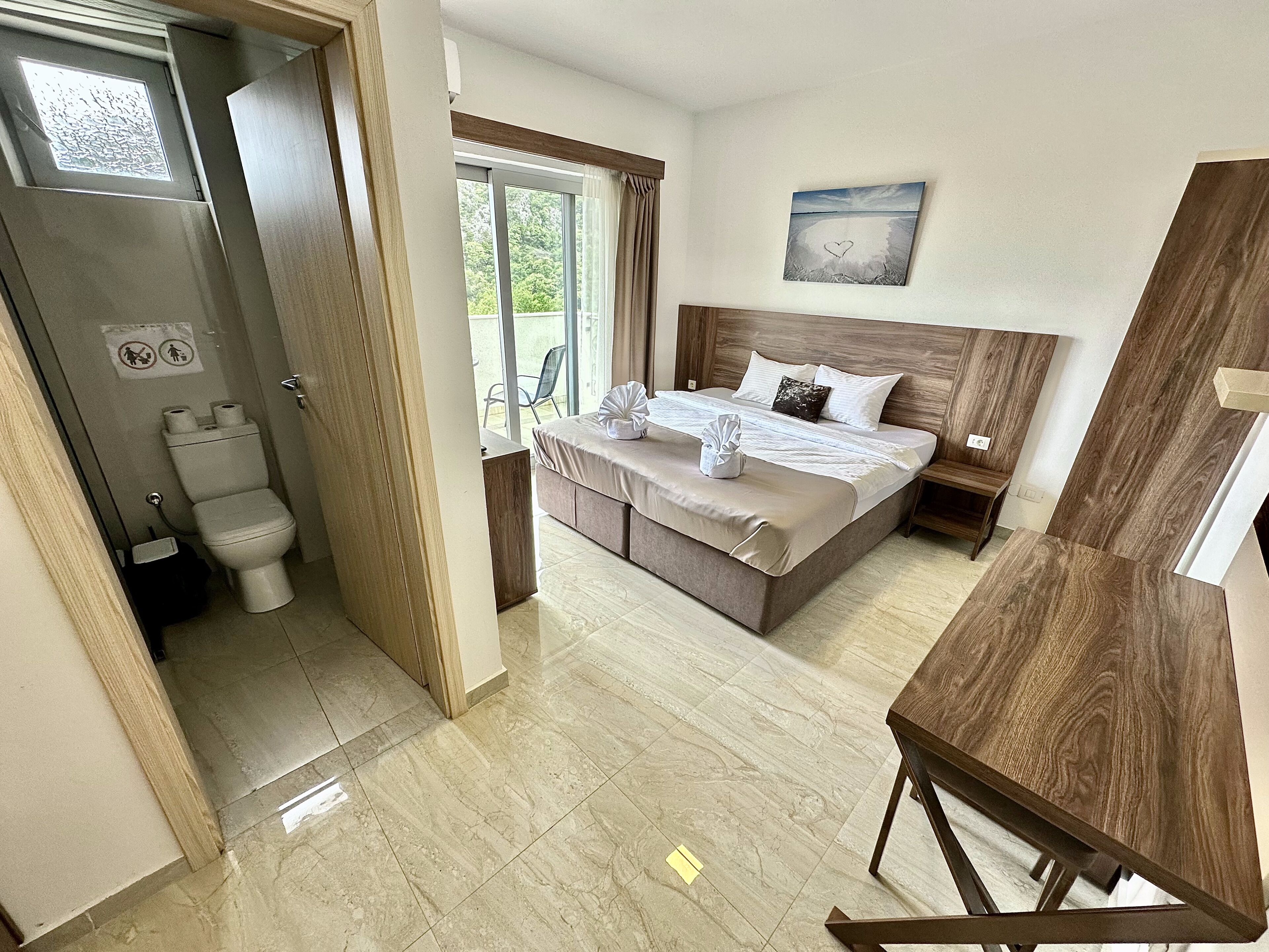 A bedroom with a bed, a wooden headboard, a nightstand, a window with curtains, and a bathroom with a toilet and sink.