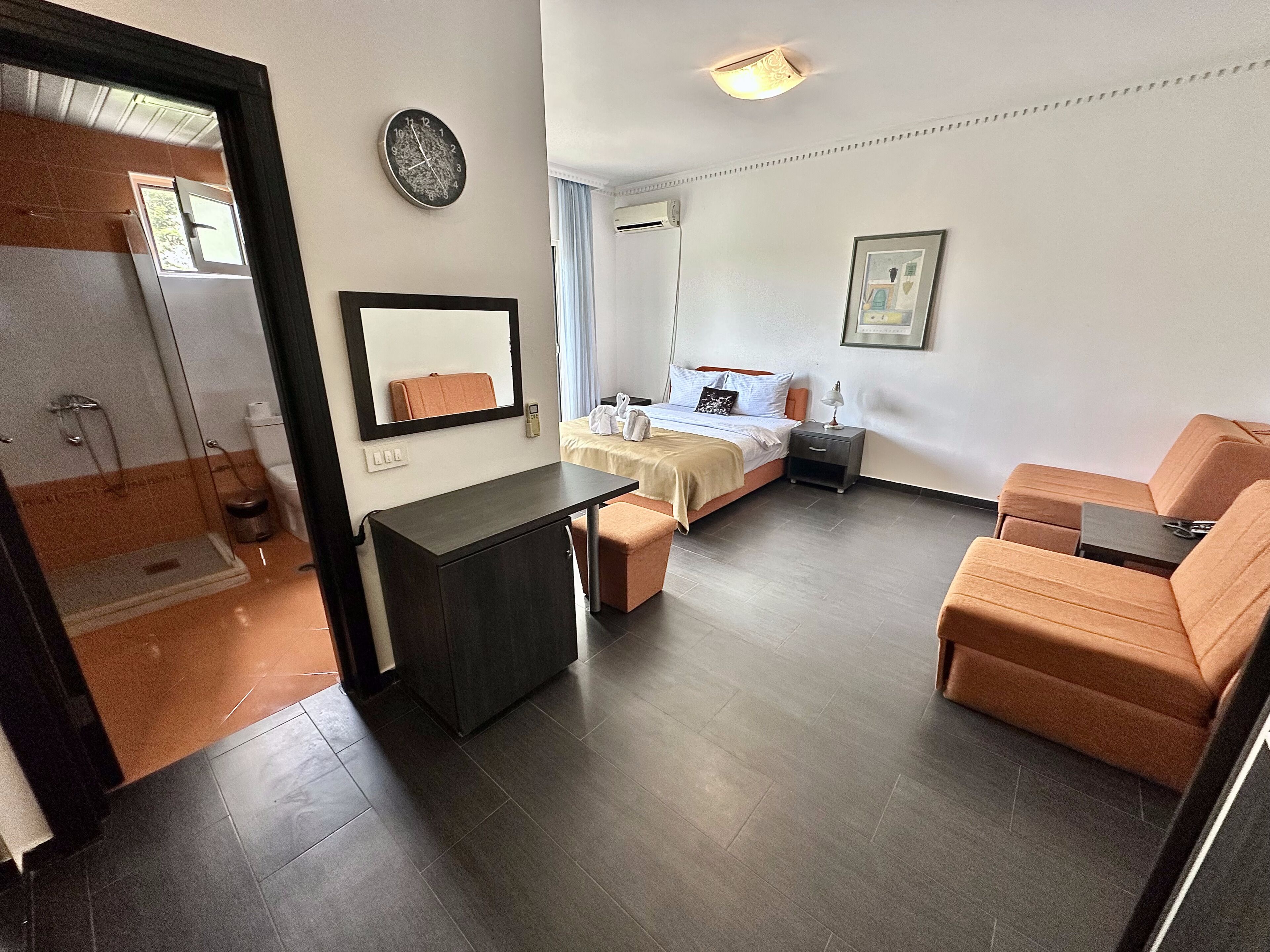 Quarto de hotel com cama, escrivaninha, cadeira, mesa pequena, mesa de cabeceira, relógio e quadro na parede.