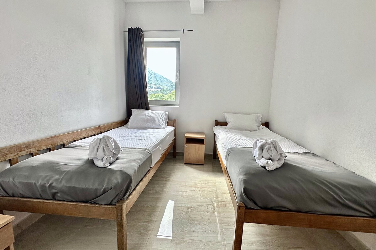 A room with two wooden beds, a window with a view of greenery, and a small wooden nightstand.