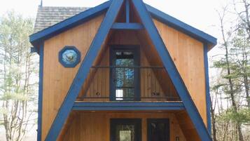 Une cabane en bois à deux étages, dotée d’un toit à forte pente, d’un balcon à l’étage supérieur et d’une porte noire.