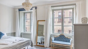 Een slaapkamer met een bed, een blauwe gestoffeerde bank, een kroonluchter en grote ramen met gordijnen.