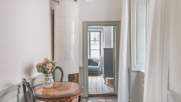 Een hal met een ronde houten tafel en stoelen, een vaas met bloemen, en zicht op een andere kamer met een bank en een raam.