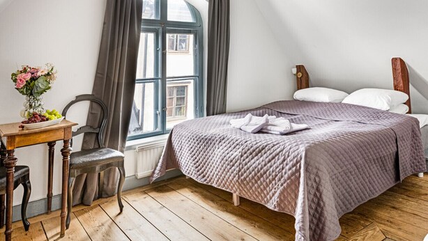 Un dormitorio con una cama, una silla de madera, una mesita de noche con un florero y una ventana con cortinas.