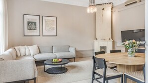 A modern living room with a grey sofa, a round wooden dining table, and a coffee table with fruit. There are framed artworks on the wall and a chandelier above.