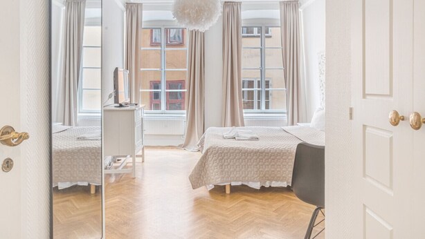 A hotel room with two beds, a desk, a chair, and a large window with curtains. - Unik lägenhet i gamla stan (Södermalm)