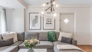 Un salon moderne avec un canapé d’angle gris, une table basse en marbre et un lustre. Des œuvres d’art encadrées ornent le mur, et une fenêtre offre une vue agréable.