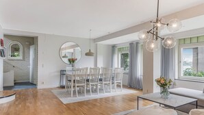 Un vaste espace de vie comprenant une table à manger, des chaises, un canapé et une table basse. Une grande fenêtre, agrémentée de rideaux, laisse entrer la lumière, et un lustre illumine l’espace. De la fenêtre, on peut admirer la verdure environnante.