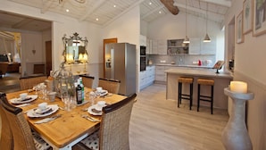 Een ruime eethoek met een houten tafel voor zes personen, omringd door rieten stoelen. Een grote kroonluchter hangt aan het plafond, en er is een moderne keuken met roestvrijstalen apparatuur zichtbaar op de achtergrond.