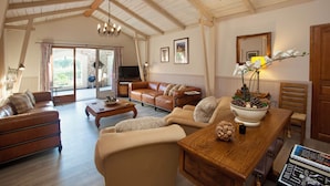 Een ruime woonkamer met een bruine leren bank, beige fauteuils, een houten salontafel en een eettafel met een bloemistdecoratie.