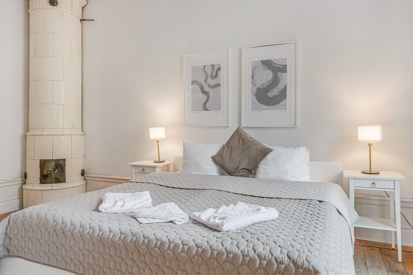 A neatly arranged bedroom with a large bed, two bedside tables, a wall-mounted fireplace, and two framed artworks.