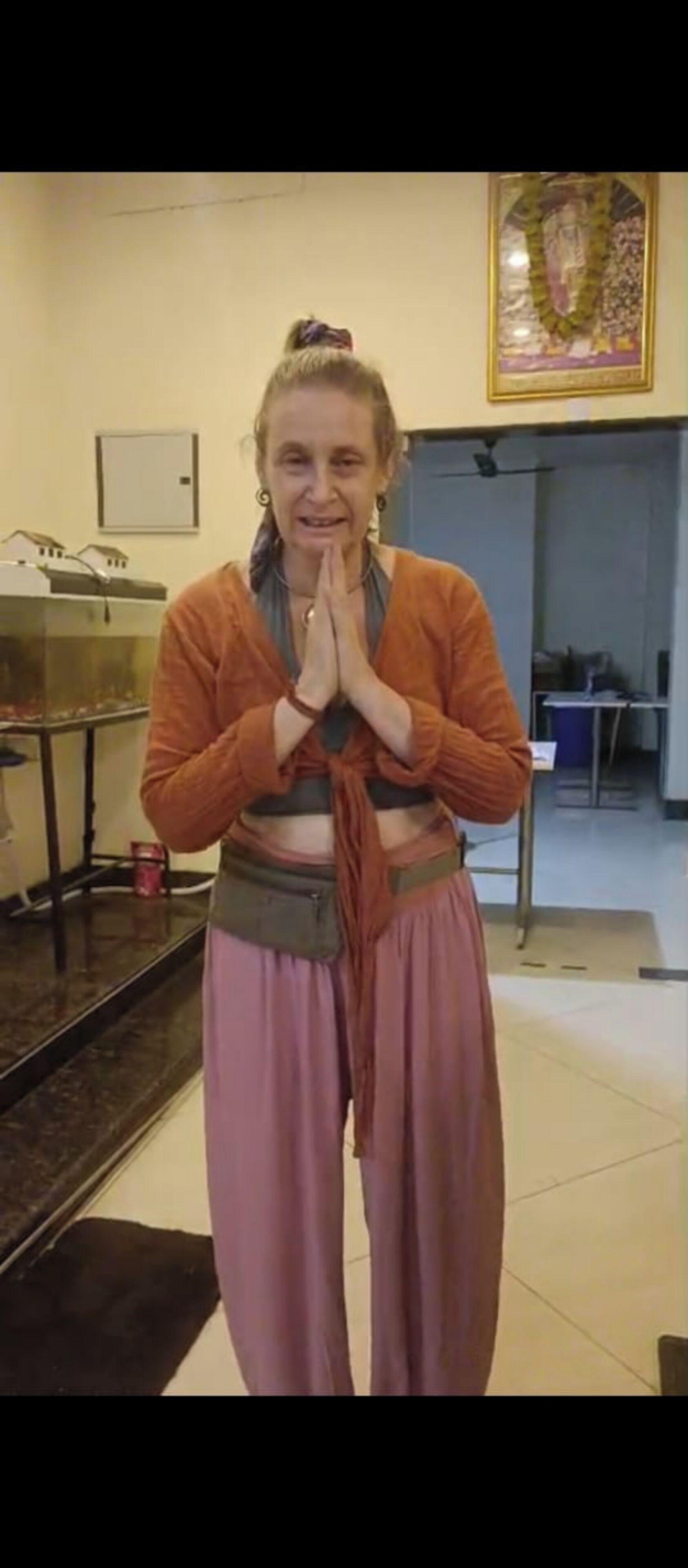 A person standing indoors with a prayer gesture, wearing an orange top and pink pants.