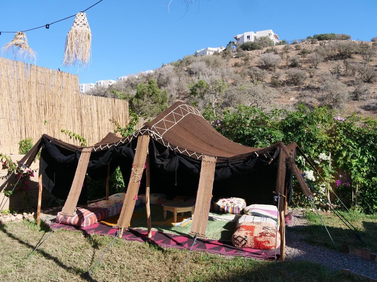 Una tienda tradicional con techo tejido y estructura de madera, instalada en un área con césped, cerca de una valla de madera y una colina al fondo.