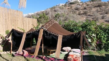 Una tienda tradicional con techo tejido y estructura de madera, instalada en un área con césped, cerca de una valla de madera y una colina al fondo.