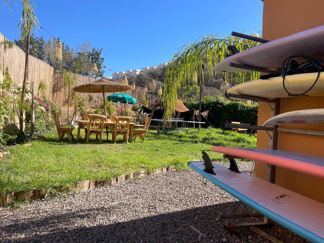 Un patio con mesa y sillas, un paraguas pequeño y una tabla de surf apoyada en una pared.