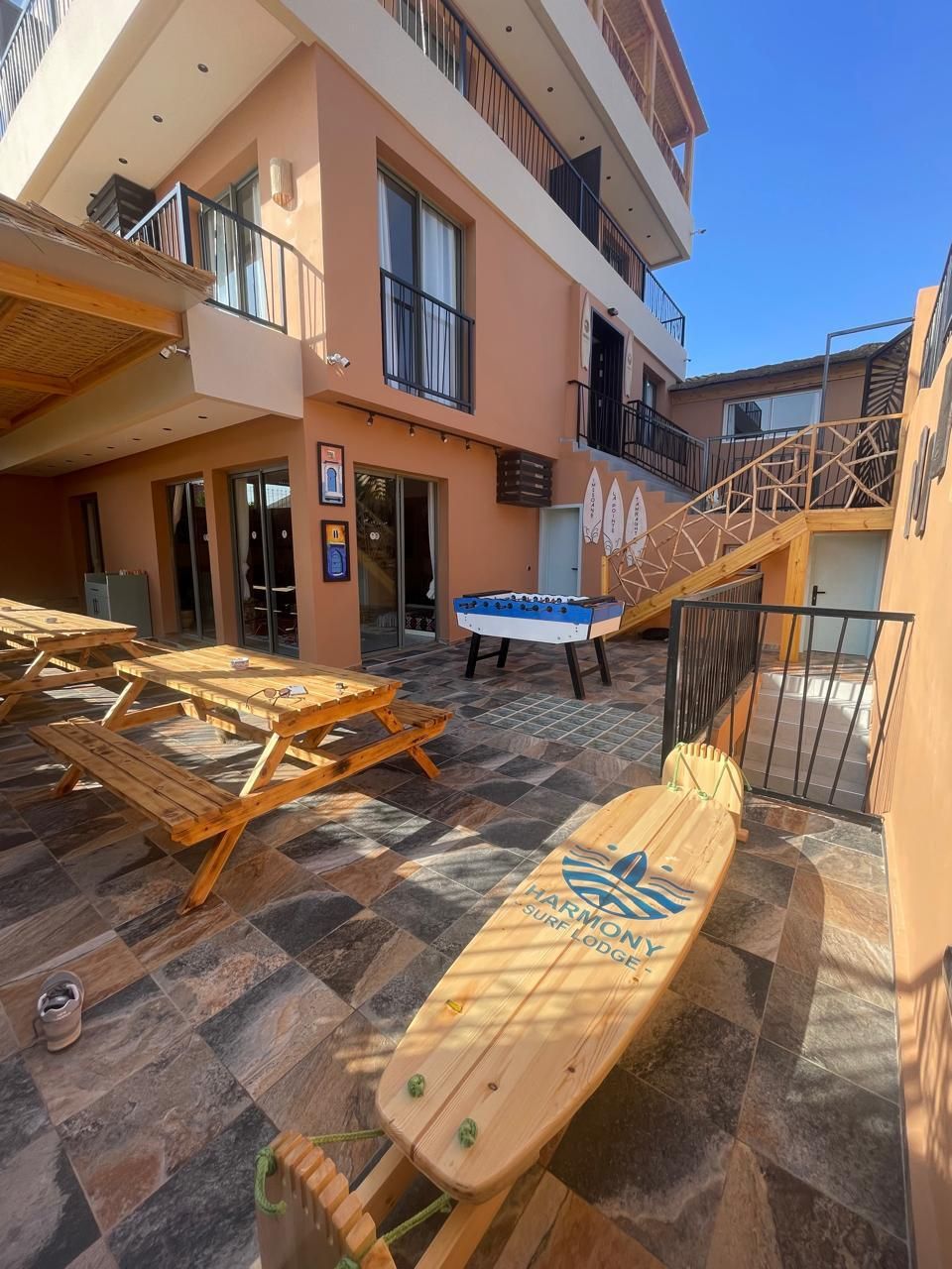 Een zwembadgedeelte met houten ligbedden en een paddleboard, een houten terras, en een gebouw met balkons en een trap.