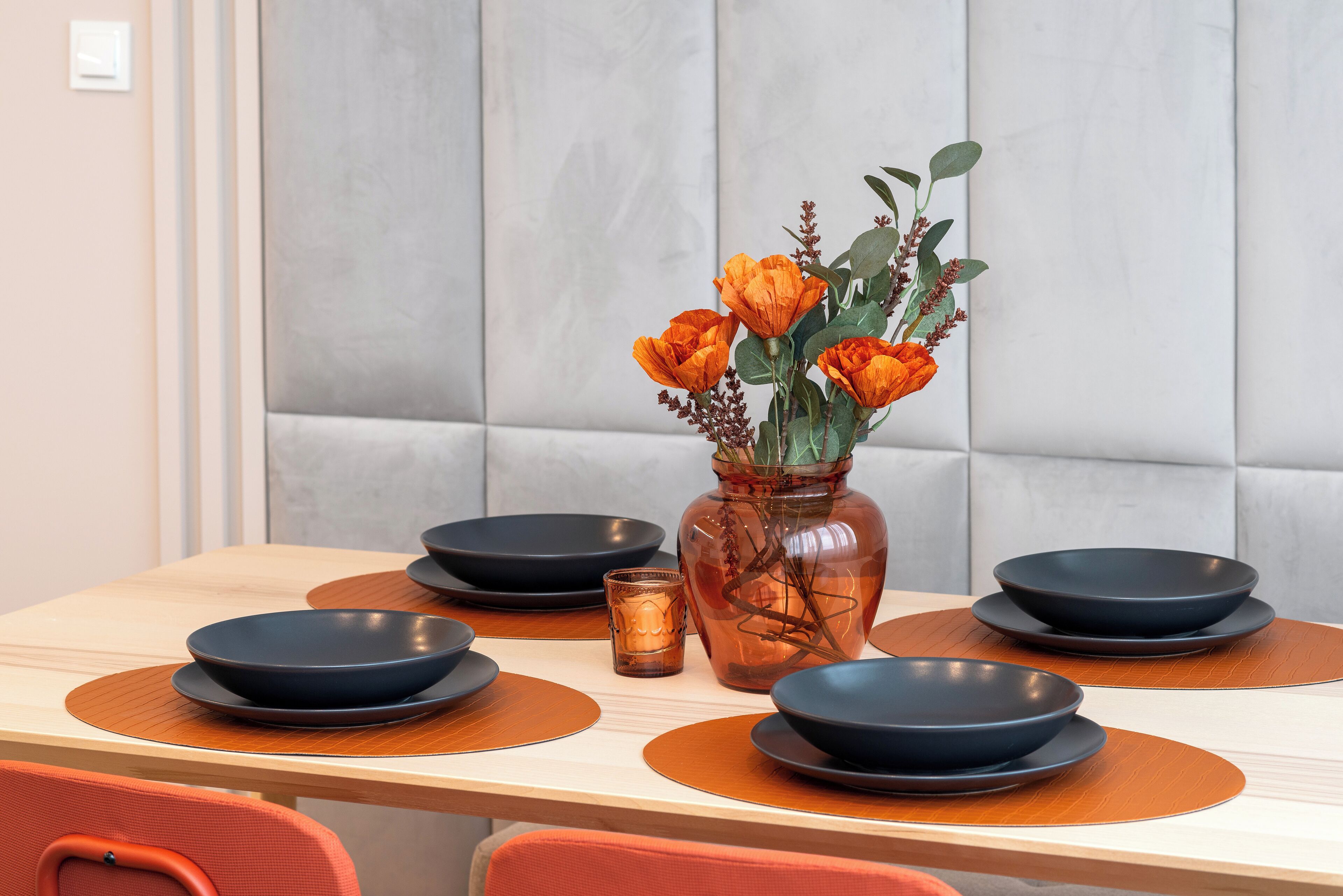 Ein Esstisch mit schwarzen Tellern, einer klaren Glasvase mit orangefarbenen Blumen und einer kleinen Kerze.