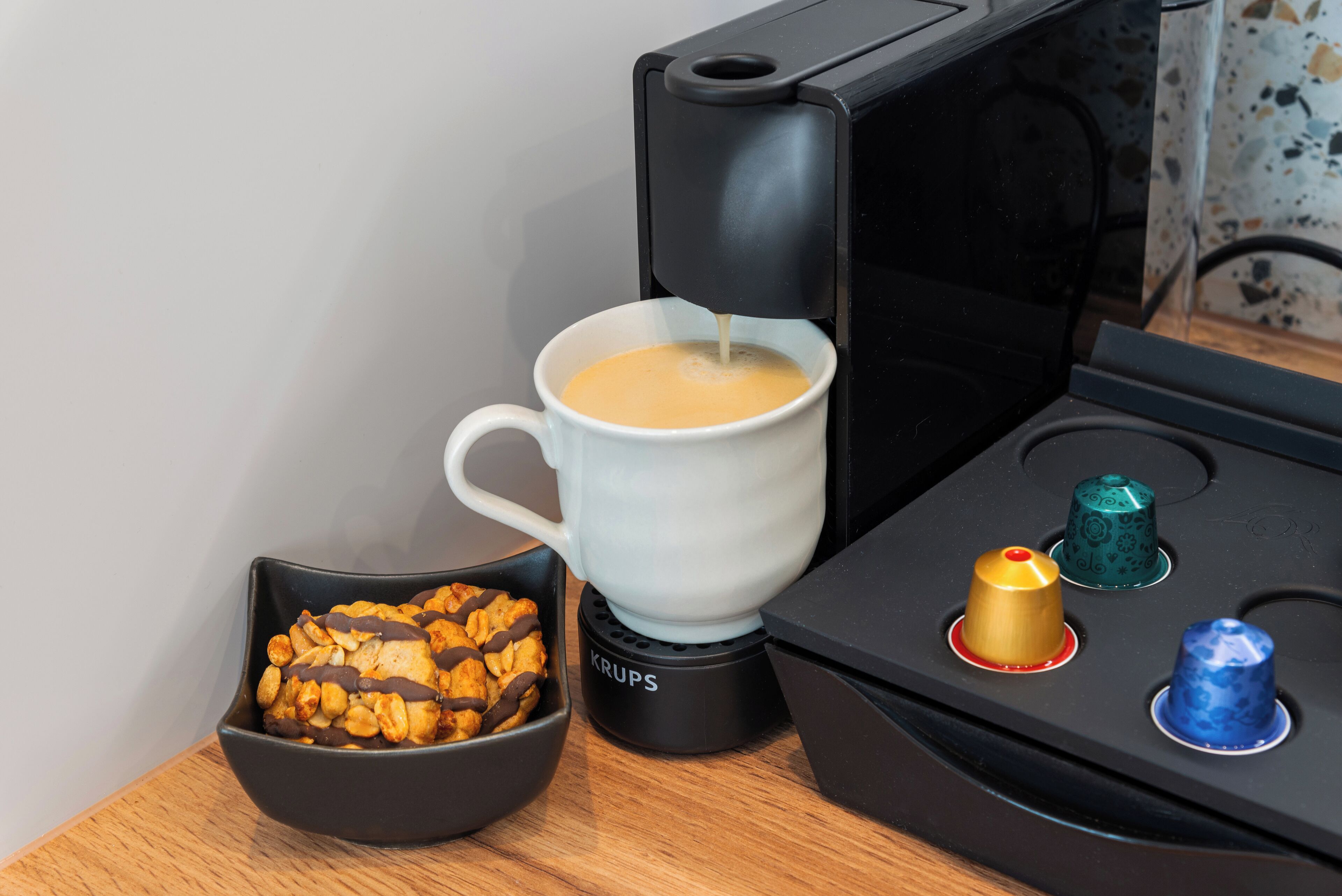 A coffee machine dispensing coffee into a cup, a bowl of cookies, and a Krups coffee maker on a wooden surface.