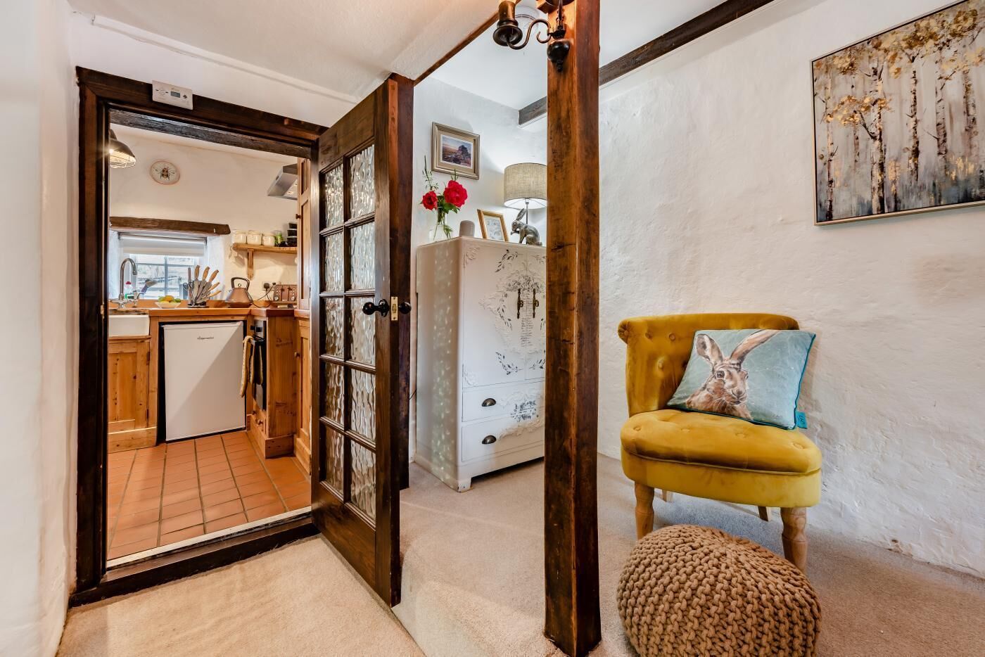 A cozy interior with a wooden-framed door leading to a kitchen, a yellow armchair with a cushion, and a white cabinet with artwork.