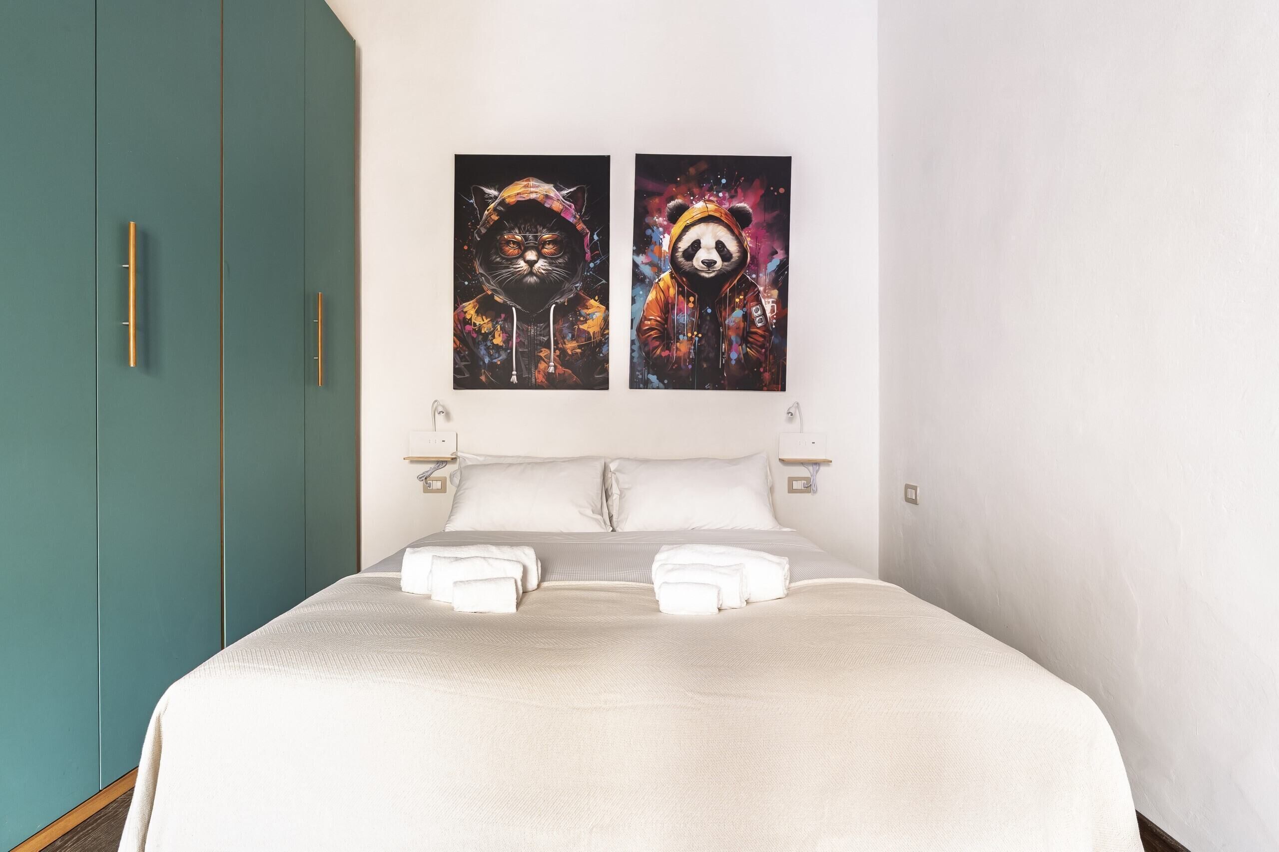 A hotel room with a teal wardrobe, a bed with white linens, and two wall art pieces depicting animals in colorful attire.