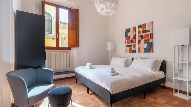 Un dormitorio con cama, una silla, una mesita, un radiador, un ventanal y un candelabro.
