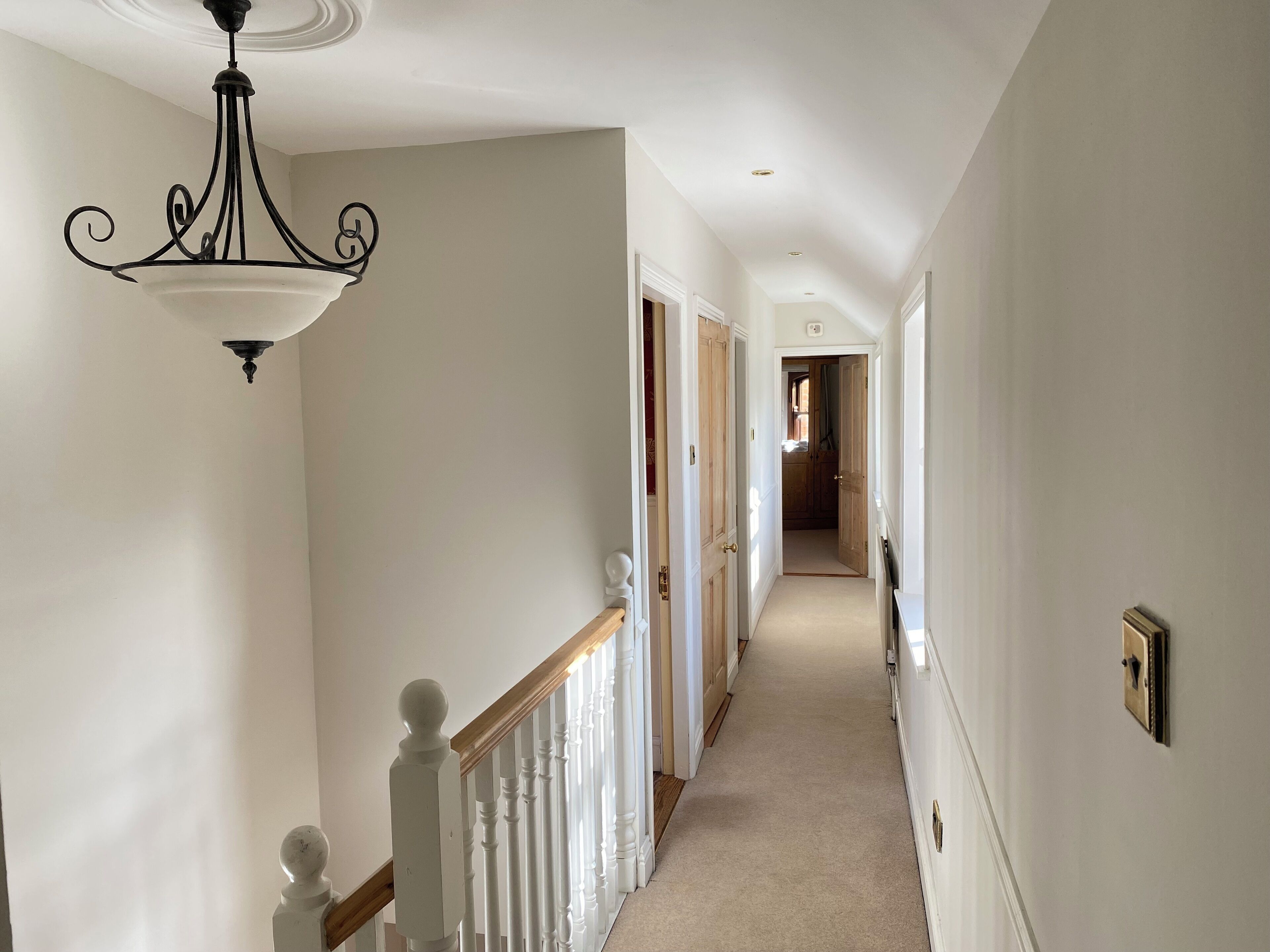Un couloir étroit et bien éclairé, aux murs blancs, avec un escalier en bois et un lustre.
