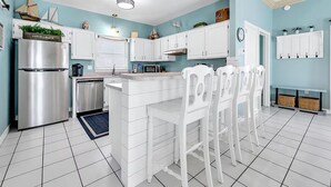 A modern kitchen with white cabinetry, a stainless steel refrigerator, and a central island with bar stools. - 🌊Private Pool-Walk to Beach-4 Bikes-Tennis Court-Tropical Dreams🌊 (Destin)