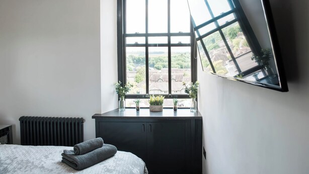 Une chambre avec une grande fenêtre, un lit avec des serviettes pliées, un radiateur et une commode de couleur sombre.