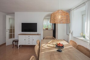 Un coin repas avec une table en bois, des chaises en osier, un téléviseur fixé au mur et un panier de fruits posé sur la table.
