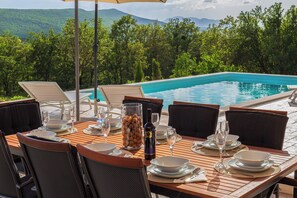 Une table à manger dressée pour six personnes, avec vue sur une piscine et des montagnes en arrière-plan.