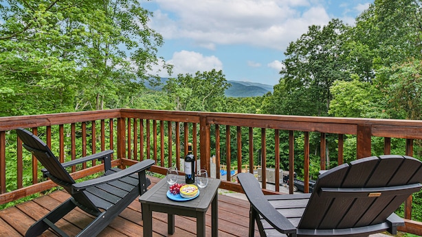 Terraza de madera con dos sillas Adirondack, una mesa con una botella y vasos, y una vista panorámica de árboles y montañas.