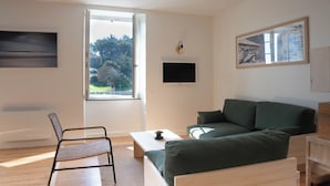 A modern living room with a green sofa, a wooden coffee table, a wicker chair, a flat-screen TV, and a large window with a view of greenery.