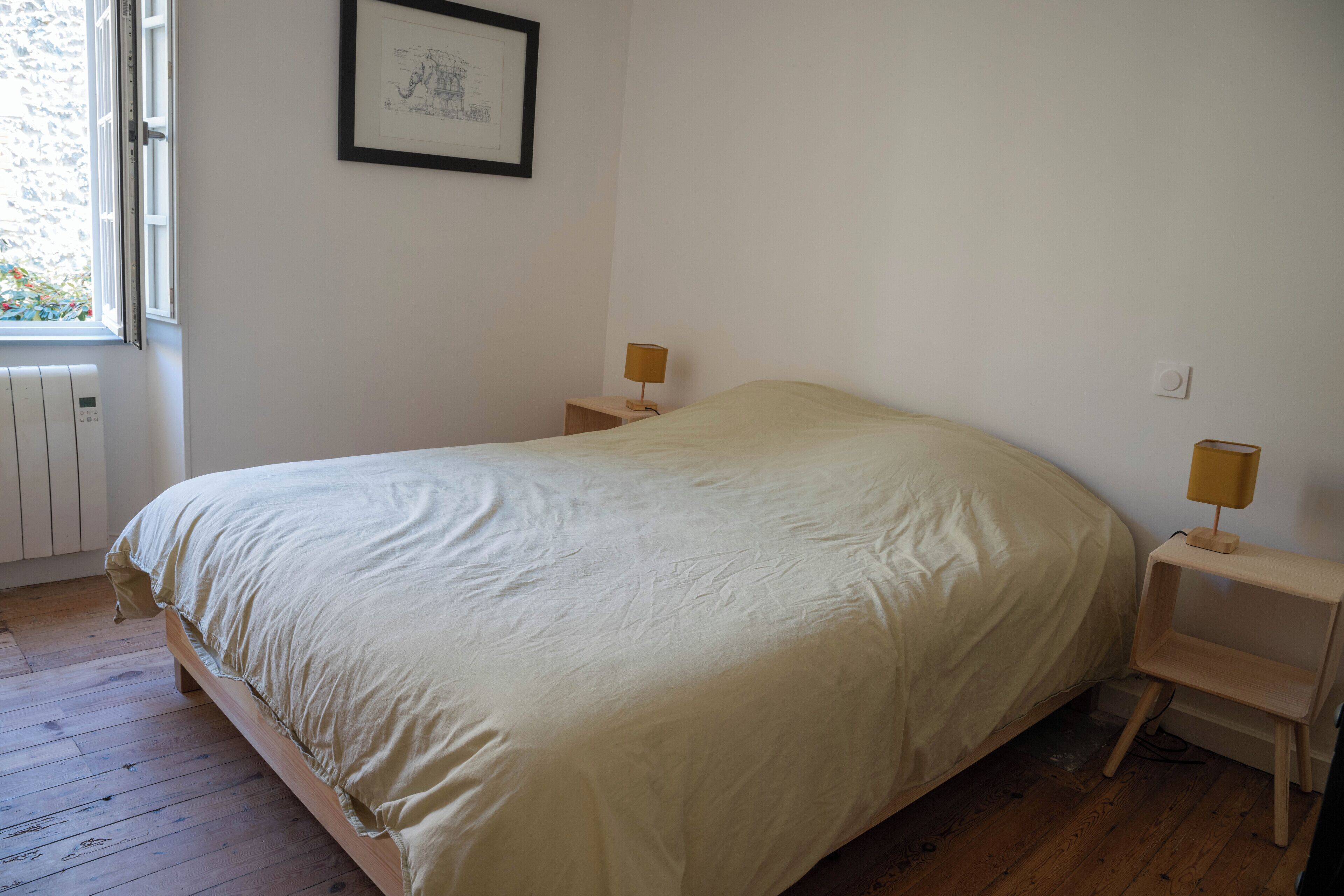 A bedroom with a large bed, two bedside tables with lamps, a window with blinds, and a framed picture on the wall.
