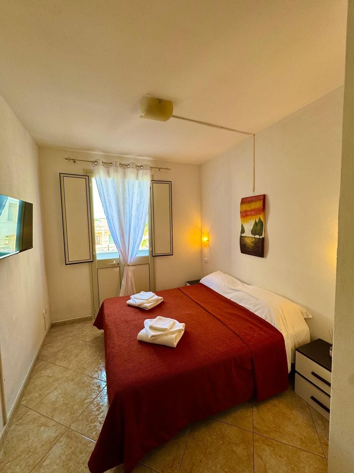 A small, clean room with a bed, a nightstand, a wall-mounted TV, and a window with curtains.