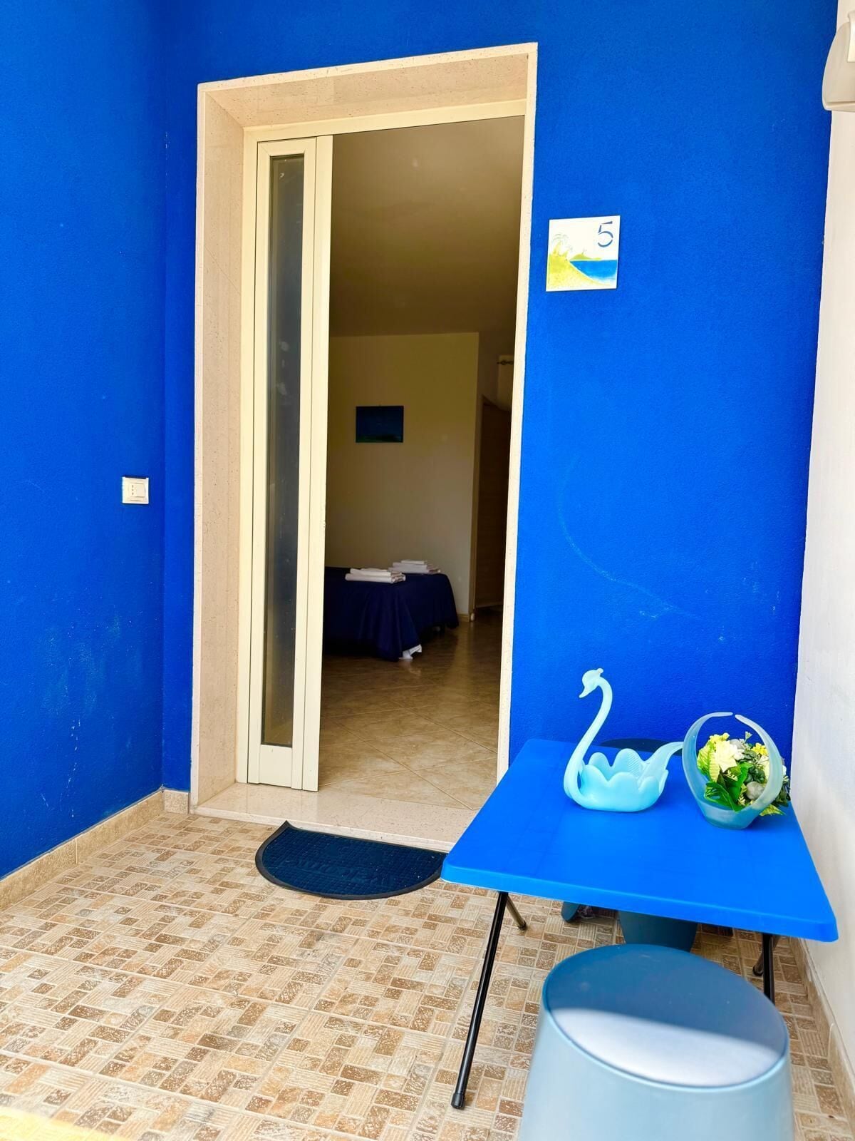 A room with blue walls, a bed with a blue bedspread, a small table with a swan-shaped container and a flower vase, and a door leading to another room.