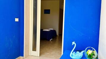 A room with blue walls, a bed with a blue bedspread, a small table with a swan-shaped container and a flower vase, and a door leading to another room.