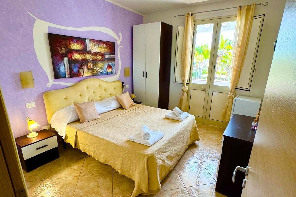 A bedroom with a bed, a wardrobe, a nightstand, a painting on the wall, and a view of the outdoors through a window.
