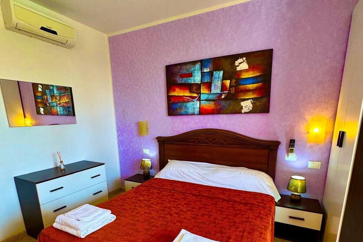 A neatly made bed with a red bedspread and two folded white towels, a wooden nightstand with a lamp, a bedside table with a lamp, a wall-mounted air conditioner, and a painting above the bed.
